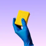 Hand in blue cleaning glove holding yellow sponge, ready for professional cleaning services.